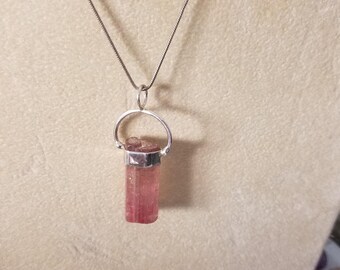 May include: A silver chain necklace with a pink and clear crystal pendant. The pendant is a long, thin, rectangular shape and is set in a silver bezel.