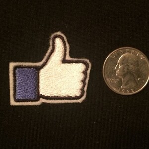 May include: Embroidered patch in the shape of a thumbs-up, with a white thumb and hand, a blue square, and a black border. A U.S. quarter coin is next to the patch. The patch is on a black background.