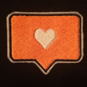 May include: An embroidered patch featuring an orange speech bubble with a white heart in the center. The patch has a black border with a white outline, set against a black fabric background. The design is simple and bold.