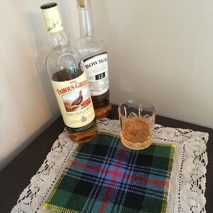 Op de afbeelding: Twee flessen Schotse whisky, een met het label "The Famous Grouse Finest Scotch Whisky" en de andere met het label "Bowmore 12", staan op een houten tafel met een glas whisky en een geruite onderzetter.