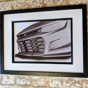 May include: A black and white photo of the front of a silver car. The car has a black grille and a black bumper. The photo is framed in a black frame.