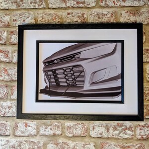 May include: A black and white photo of the front of a car, showing the grill and headlights. The car is framed in a black frame.