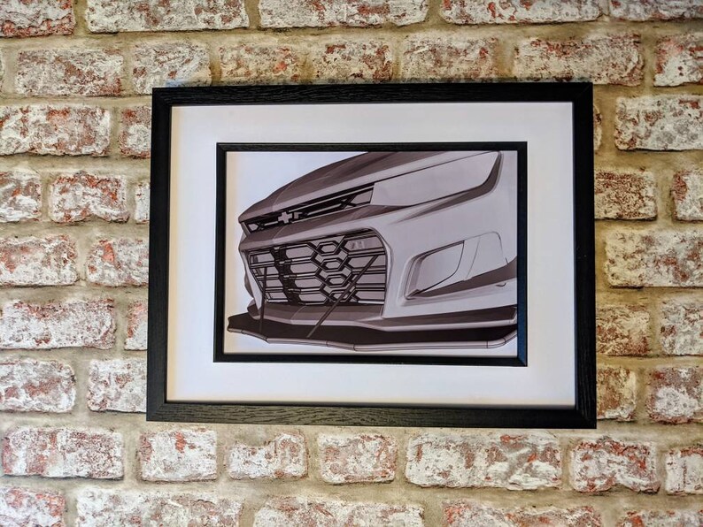 May include: A black and white photo of the front of a car, showing the grill and headlights. The car is framed in a black frame.