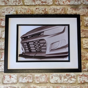 May include: A black and white photo of a car's front grille, showing the headlights, grille, and bumper. The photo is framed in a black frame.