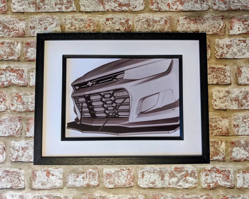 May include: A black and white photo of a car's front grille, showing the headlights, grille, and bumper. The photo is framed in a black frame.
