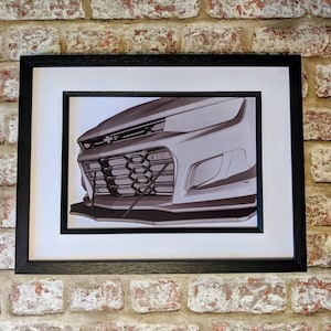 May include: A black and white photo of a car's front grille, showing the headlights, grille, and bumper. The photo is framed in a black frame.