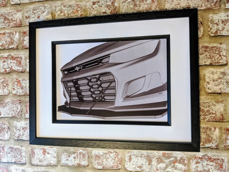 May include: A black and white photo of the front of a silver car. The car has a black grille and a black bumper. The photo is framed in a black frame.
