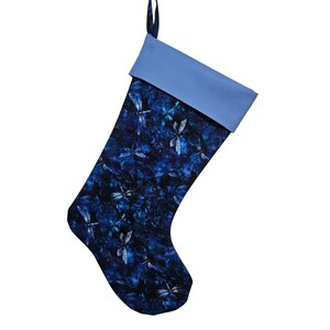 May include: A holiday stocking featuring a dark blue fabric body with a repeating pattern of light blue and white dragonflies. The stocking has a solid light blue cuff at the top and a matching fabric loop for hanging, set against a plain white background.