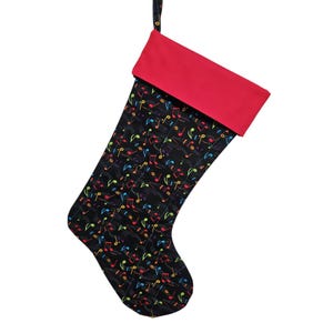 May include: A Christmas stocking featuring a black fabric body patterned with colorful musical notes in red, blue, yellow, and green. The stocking has a solid red cuff at the top and a small fabric loop for hanging, presented against a plain white background.