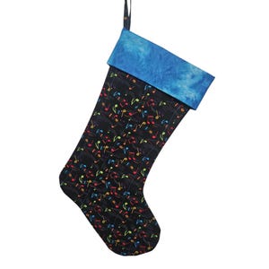 May include: A holiday stocking featuring a black fabric body patterned with colorful musical notes in red, green, yellow, and blue. The cuff is made of a textured blue fabric, and a thin black fabric loop is attached to the top for hanging.