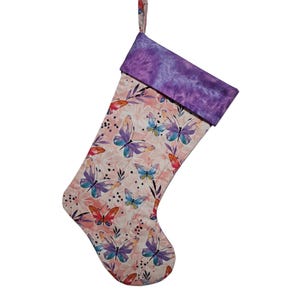 May include: A Christmas stocking featuring a light pink background with a repeating pattern of colorful butterflies in shades of purple, blue, and orange, accented by small black dots and floral outlines, finished with a solid purple cuff.