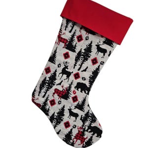 Peut inclure: Une chaussette de Noël avec un revers et une boucle de suspension rouges. Le corps de la chaussette est orné d'un motif d'arbres noirs, de cerfs et d'ours sur fond blanc. Des cerfs et des losanges rouges à carreaux ajoutent de la couleur.