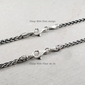 May include: Two sterling silver wheat chains featuring lobster claw clasps. The top chain has a Clasp With Dots design, while the bottom chain features a Clasp With Fleur de lis. Both clasps are stamped with 925 to indicate silver purity.
