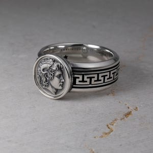 May include: A silver ring featuring a circular coin-style face with a relief profile of a figure with ram horns. The band is decorated with a black engraved Greek key pattern around the circumference and includes small star details on the inner band.