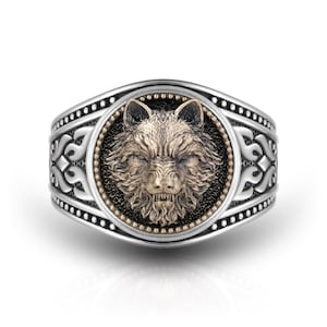 May include: A silver ring featuring a central circular medallion with a detailed bronze-toned wolf head relief. The wolf is set against a dark, textured background surrounded by a beaded border. The band displays intricate, symmetrical scrollwork.