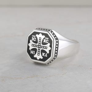 May include: A silver signet ring featuring an octagonal face with a black inlay. The center displays a white cross with the Greek letters IC XC NI KA arranged in the four quadrants. The border of the octagonal face is detailed with small beads.