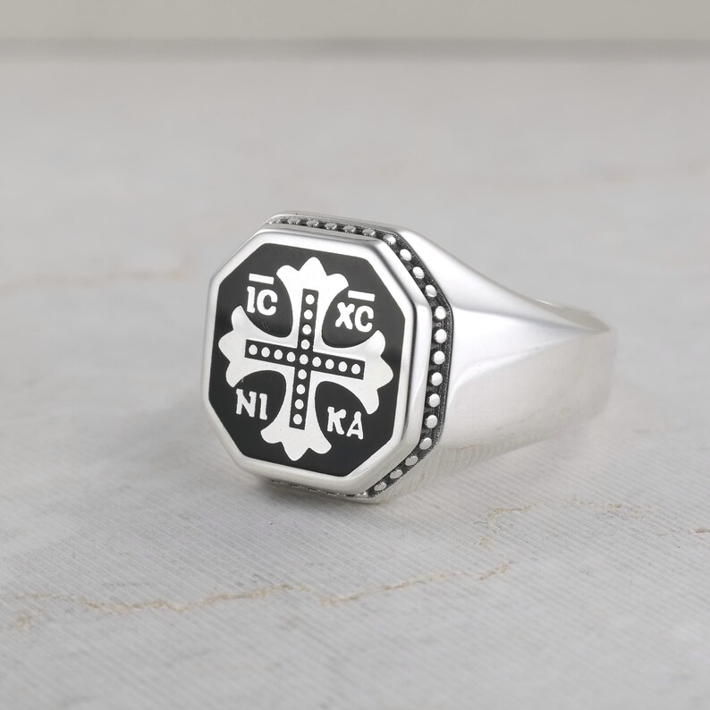 May include: A silver signet ring featuring an octagonal face with a black inlay. The center displays a white cross with the Greek letters IC XC NI KA arranged in the four quadrants. The border of the octagonal face is detailed with small beads.