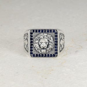 Puede incluir: Un anillo de tono plateado con una cara cuadrada que presenta un relieve detallado de una cabeza de león en el centro. El león está rodeado por un borde de pequeñas piedras azul oscuro. Los lados del anillo muestran intrincados grabados.
