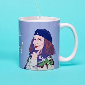 Peut inclure: Une tasse en céramique blanche avec un fond bleu et une illustration de dessin animé d'une femme aux cheveux roux portant une écharpe verte et blanche et un béret noir. Le texte "Darling, you're looking smashing!" est imprimé sur la tasse.