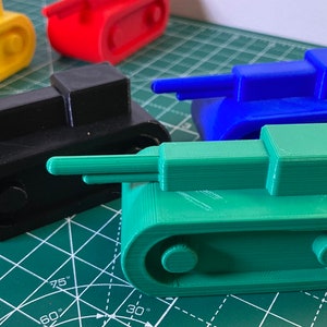 Può includere: Una collezione di carri armati giocattolo colorati in giallo, rosso, blu, nero e verde. Ogni carro armato ha un corpo rettangolare, una torretta e una canna lunga. I giocattoli sono disposti su un tappetino da taglio verde.