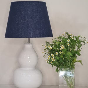 May include: A white ceramic table lamp with a blue fabric lampshade. The lamp has a gourd-shaped base. A vase with white flowers is in the background.