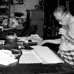 May include: A man sits at a table, working on a project. He is wearing a plaid shirt and has a focused expression on his face. The table is covered in fabric and tools, suggesting a craft or sewing project.