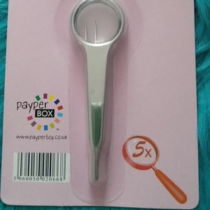 Puede incluir: Pinzas de precisión con lupa plateadas con una lente de aumento de 5x. El embalaje es rosa y presenta el logotipo de Payper Box y la dirección del sitio web. El código de barras es 5060050 020668.