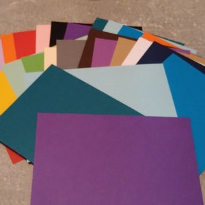 Puede incluir: Un abanico de hojas de papel de colores en varios tonos de azul, verde, morado, rojo, naranja, amarillo, marrón y gris. El papel está dispuesto en un semicírculo, con la hoja más grande en el centro.