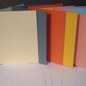 Peut inclure: Une pile de cartes de v&oelig;ux vierges de diff&eacute;rentes couleurs, dont cr&egrave;me, bleu, orange, jaune, rose et bleu clair. Les cartes sont dispos&eacute;es en &eacute;ventail, avec la carte cr&egrave;me &agrave; l'avant.