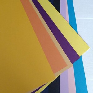 May include: A stack of colorful paper sheets in various shades of yellow, orange, purple, blue, and pink. The sheets are arranged in a fan shape, with the yellow sheet at the top and the pink sheet at the bottom.
