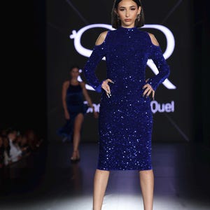 May include: A woman in a royal blue, sequined, knee-length dress with long sleeves and shoulder cutouts. She is wearing black high heels. The background features a logo and the word "Quel".