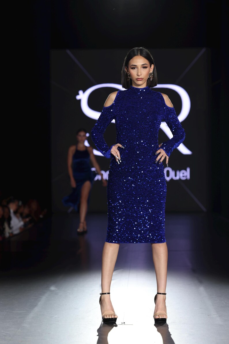 May include: A woman in a royal blue, sequined, knee-length dress with long sleeves and shoulder cutouts. She is wearing black high heels. The background features a logo and the word "Quel".