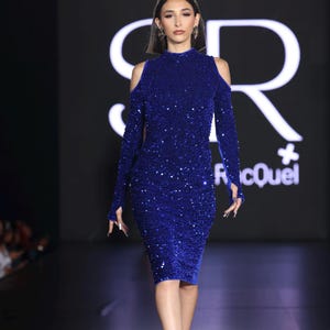 May include: A woman in a royal blue, sequined, long-sleeved dress with shoulder cutouts. The dress is knee-length and has a high neckline. She is wearing black heels. The background is dark with white text.