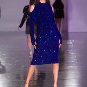 May include: A long-sleeved, knee-length, royal blue dress with a high neckline and shoulder cutouts. The dress is covered in sequins and is paired with gold heels. The model is on a runway.