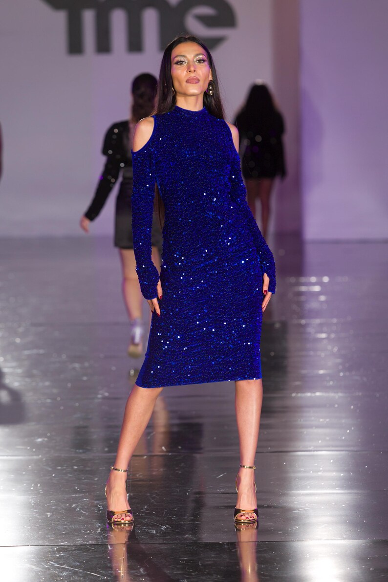 May include: A long-sleeved, knee-length, royal blue dress with a high neckline and shoulder cutouts. The dress is covered in sequins and is paired with gold heels. The model is on a runway.
