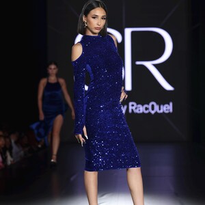 May include: A model in a royal blue sequined dress with long sleeves and shoulder cutouts. The dress is knee-length and paired with clear strap heels. The background features a fashion show runway with the brand name "y RacQuel".