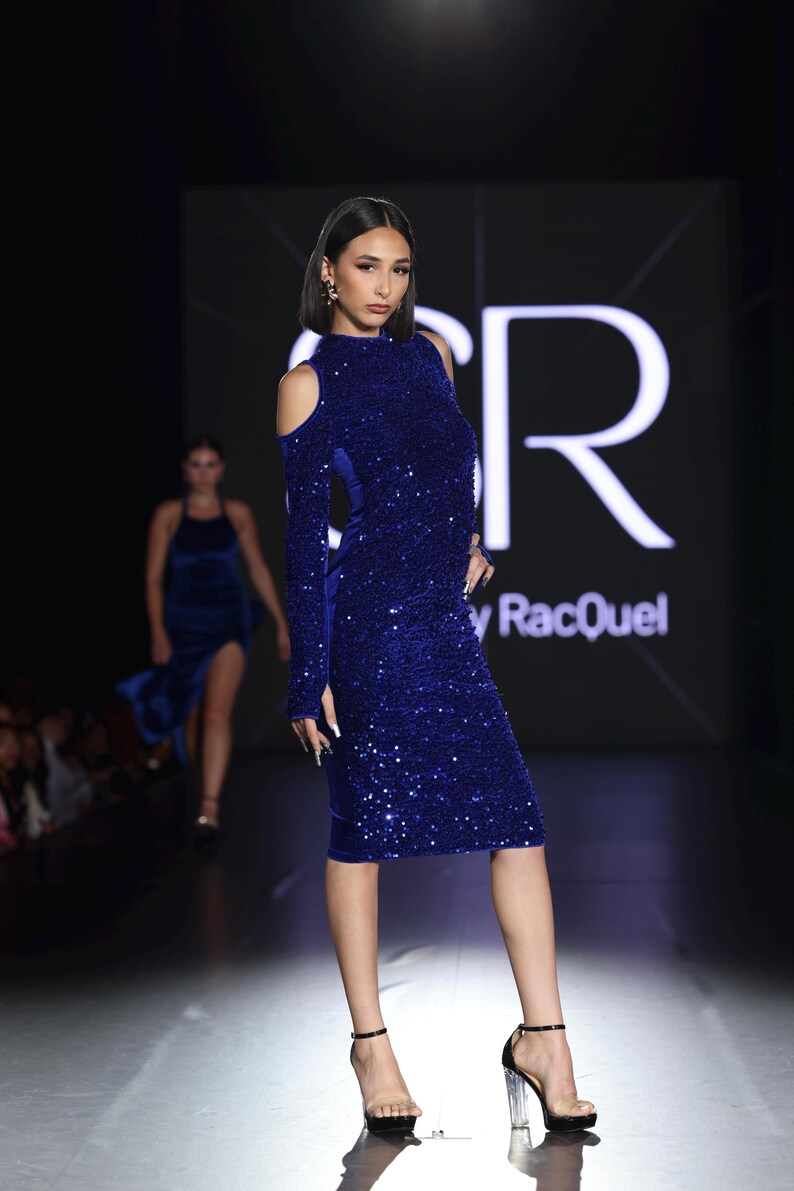 May include: A model in a royal blue sequined dress with long sleeves and shoulder cutouts. The dress is knee-length and paired with clear strap heels. The background features a fashion show runway with the brand name "y RacQuel".