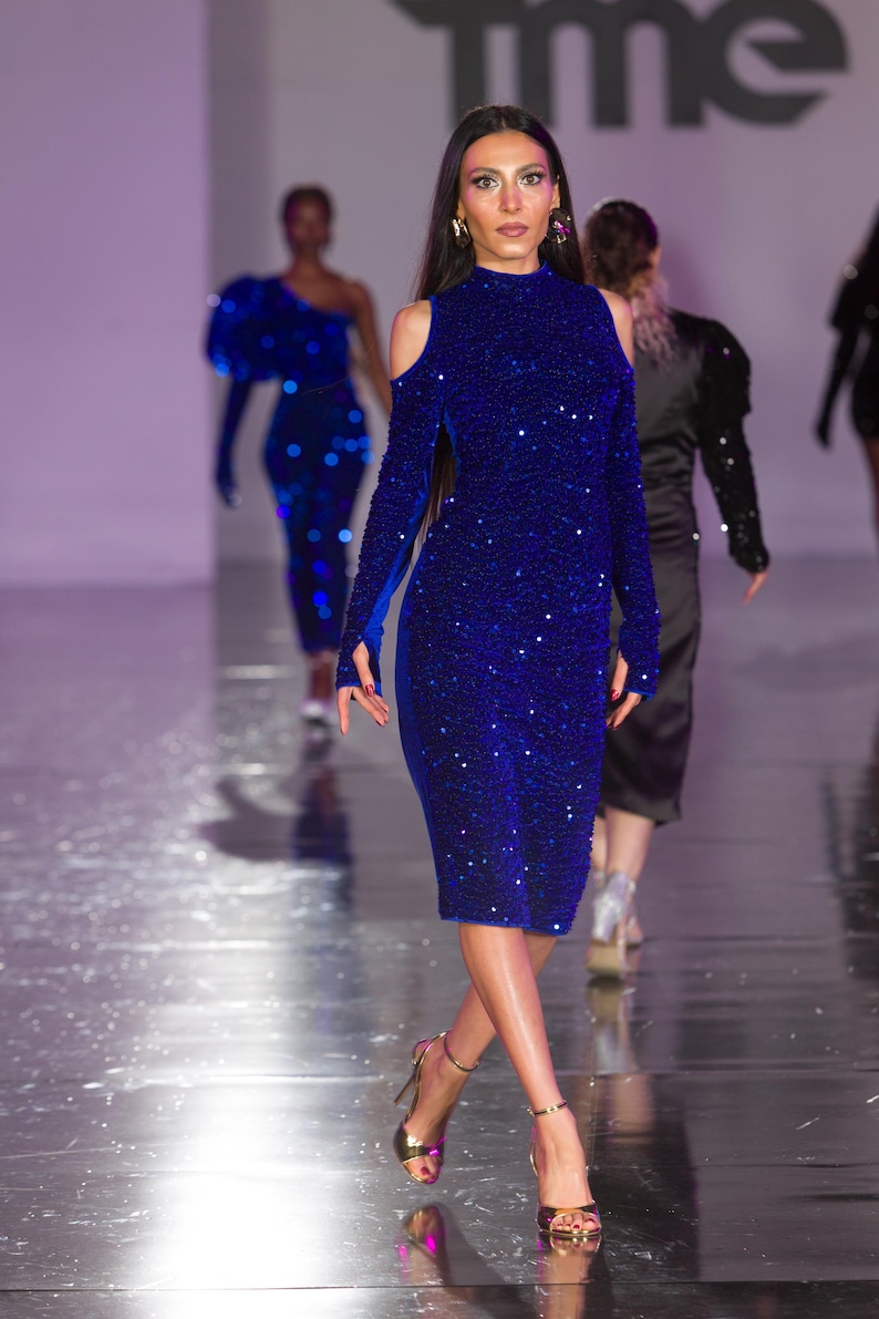 May include: A model in a royal blue, knee-length dress with long sleeves and shoulder cutouts walks on a runway. The dress is covered in sequins. She wears gold heels. Other models are in the background.