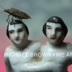 Könnte beinhalten: Zwei Porzellanfiguren von Menschen mit schwarzen Haaren, eine mit einem Käfer auf dem Kopf. Die Figuren sind weiß mit bemalten Details und sind so gestellt, als würden sie ringen.