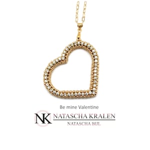 May include: Gold heart-shaped pendant necklace with clear crystal accents. The necklace is on a gold chain and has the text "Be mine Valentine" and "NATASCHA KRALEN NATASCHA BIJL" on the bottom.