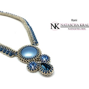 May include: A silver and blue beaded necklace with a large blue pendant featuring a round cabochon stone and smaller blue stones. The necklace is designed by Natascha Kralen, Rani.