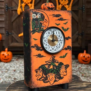 May include: A rectangular orange wooden clock featuring a black circular clock face with white numbers. The surface displays illustrations of three witches, an owl perched on a crescent moon, bats, and clouds. A small orange pumpkin sits on top.