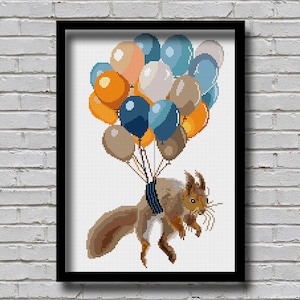 May include: A framed cross-stitch artwork shows a brown squirrel carried by a bunch of colourful balloons. The balloons are in shades of orange, blue, tan, and white. The artwork is set against a white background and framed in black.