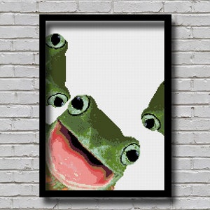 May include: A green frog with large eyes and a wide open mouth peeks out from behind a white frame. The frog's tongue is visible and its eyes are looking directly at the viewer.