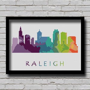 May include: A framed art print featuring a colorful, pixelated skyline of Raleigh, North Carolina. The city's silhouette is rendered in shades of purple, blue, green, and orange. The word "RALEIGH" is printed below the skyline in a matching color gradient.