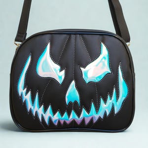 May include: A black crossbody bag featuring a stitched pumpkin face design. The eyes, nose, and jagged mouth are made of an iridescent, holographic material that reflects blue and purple tones against the matte black surface of the bag.