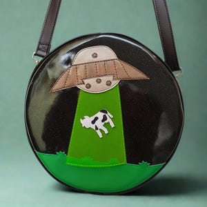 May include: Round black crossbody bag with a glittery finish, featuring a UFO design with a cow being abducted. The UFO is brown and beige, with a green beam shining down on a black and white cow. The bag has a brown strap.