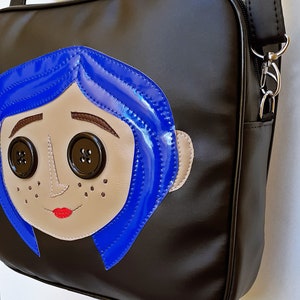 May include: Black shoulder bag featuring a cartoon face with blue hair, button eyes, and a red mouth. The bag has a long strap and a zipper closure. The cartoon face is made of tan and blue leather-like material.