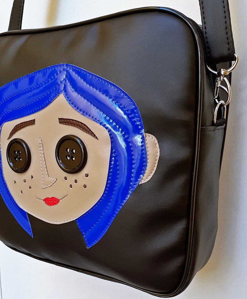 May include: Black shoulder bag featuring a cartoon face with blue hair, button eyes, and a red mouth. The bag has a long strap and a zipper closure. The cartoon face is made of tan and blue leather-like material.