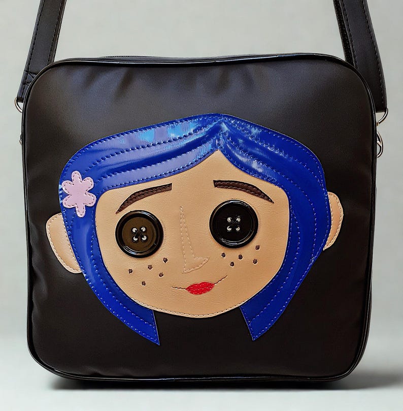 May include: Black messenger bag featuring a stylized face with blue hair, button eyes, and a pink flower. The face is a light tan color with red lips and freckles. The bag has a black adjustable strap.